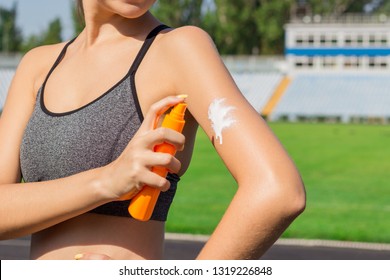 Young female runner standing and putting sun lotion on hand. Girl using sunscream before sport running exercise on summer morning. Sports and healthy concept.