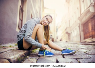 Young female runner is tying her running shoes on tiled pavement in old city center