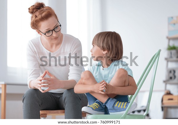 Young female school psychologist\
having serious conversation with smart little boy at her\
office
