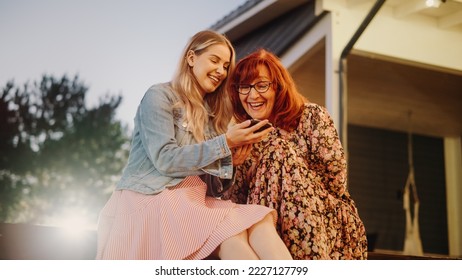 Young Female Showing Family Photos and Videos to Her Grandparent. Relatives Sitting Outdoors on a Porch of the House on a Warm Summer Day, Using Mobile Phone to Look Up Info on Internet. - Powered by Shutterstock