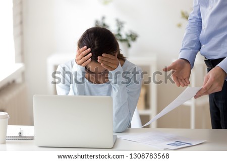 Young female sitting at workplace desk putting her head down, lack working experience employee feels guilty and desperate, company executive manager accusing office worker showing mistakes in report