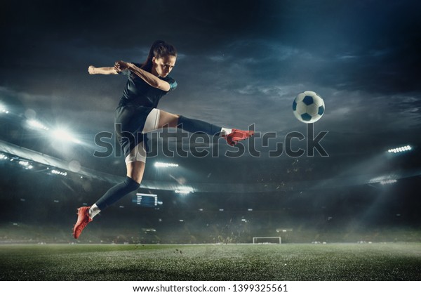 Young female soccer or football player with long\
hair in sportwear and boots kicking ball for the goal in jump at\
the stadium. Concept of healthy lifestyle, professional sport,\
hobby, motion, movement
