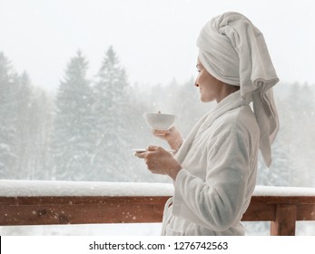 Young Female Standing After Taking A Shower In The Morning On Balcony Of The  Hotel In Winter. Holding A Cup Of Coffee Or Tea In Her Hands. Looking Outside Nature Forest And Mountain Under The Snow