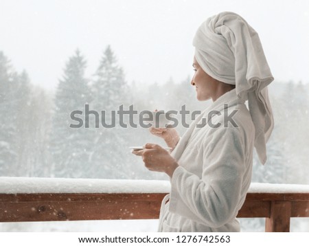 Young female standing after taking a shower in the morning on balcony of the  hotel in winter. holding a cup of coffee or tea in her hands. Looking outside nature forest and Mountain under the snow
