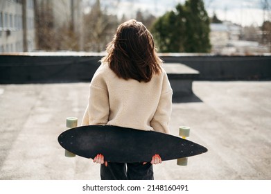 Young female standing back with sport skateboard on windy rooftop. Woman with dark hair in outwear clothes enjoying street activity and beautiful view from high building.