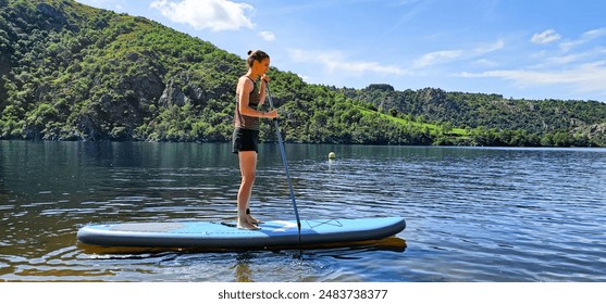 Young female standing on SUP board under blue sky on beautiful lake with amazing green tree covered hills in background; slim woman in swimming costume shorts and top on a stand up paddleboard in sun
 - Powered by Shutterstock