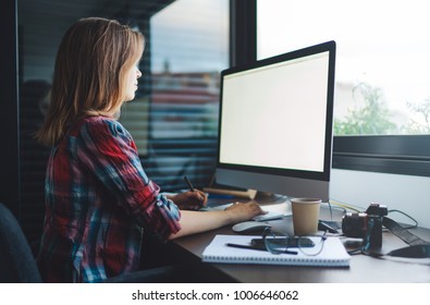 a young female stock photographer wearing in casual clothes adds photos to her account on the Photobank while sitting in a furnished office with modern technology. Blank screen with copy space