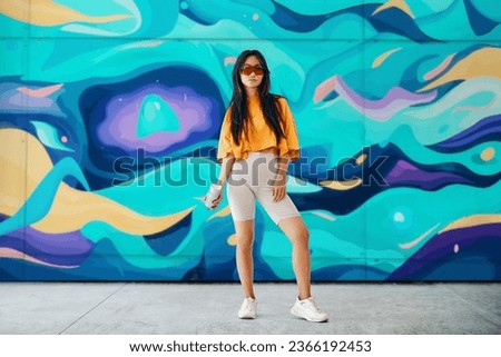 Young female street artist with spray paint can in hand looking to camera over her graffiti wall paintings background. Street art concept. Full length portrait