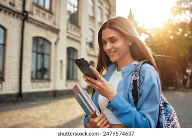 A young female student enjoys modern academic life on campus, using a smartphone while carrying books, symbolizing a vibrant youth culture of learning and studying in a university setting - Powered by Shutterstock