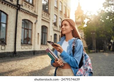 A young female student enjoys modern academic life on campus, using a smartphone while carrying books, symbolizing a vibrant youth culture of learning and studying in a university setting - Powered by Shutterstock