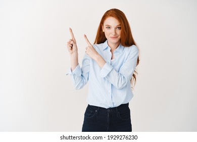 Young Female Student With Ginger Hair And Blue Eyes Showing Company Logo, Pointing Fingers Upper Left Corner And Smiling At Camera, White Background.