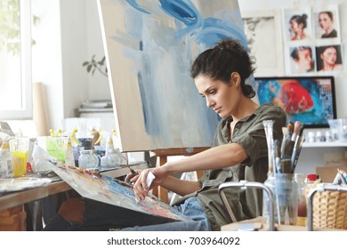 Young female student having classes at art studio, learning how to draw landscapes, trying to mix different watercolors on cardboard. Concentrated woman with dark hair, dressed casually, painting - Powered by Shutterstock