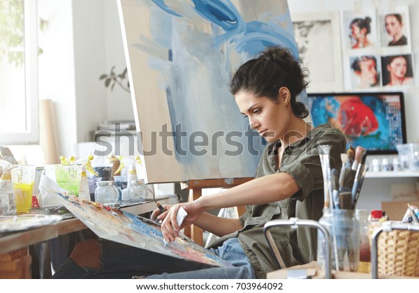 Young female student having classes at art
studio, learning how to draw landscapes, trying to mix different
watercolors on cardboard. Concentrated woman with dark hair,
dressed casually,
painting