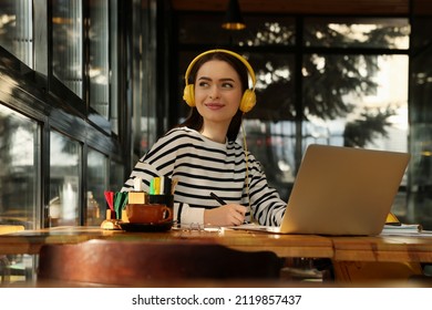 Young Female Student With Laptop And Headphones Studying At Table In Cafe