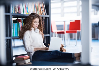 Young female student in library, focusing on final project, presentation. Sitting on floor with laptop on knees. University student preparing for final exam. - Powered by Shutterstock