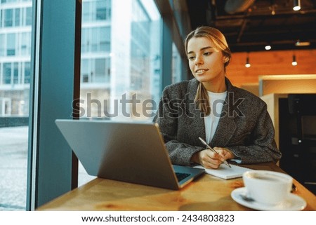 Young female student is making notes and browsing laptop sitting in cafe near window 