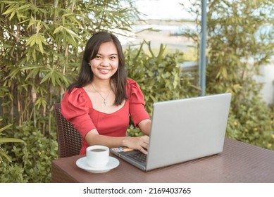 A young female student in a red blouse typing on a computer. Working on her thesis or freelance project. Outdoor scene at an al fresco cafe. - Shutterstock ID 2169403765