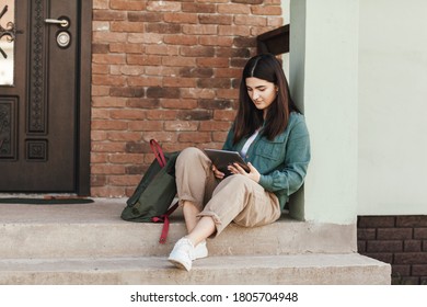 Young Female Student Sitting On Steps And Studying With Tablet In College Or Private School, Technology Teaching Concept.