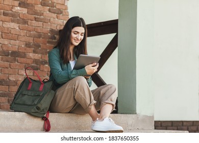 Young Female Student Sitting On Steps And Studying With Tablet In College Or Private School, Technology Teaching Concept.Technology, Education, Leisure Concept