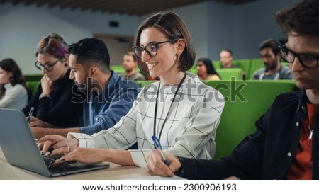 Young Female Student Studying in University with Diverse Multiethnic Classmates. She is Focused and Using a Laptop Computer. Patiently Listening to Professor's Lecture and Taking Notes
