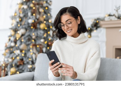 A young female student is texting on the phone, reading the news. He sits on the sofa near the Christmas tree and looks at the screen with a smile. - Powered by Shutterstock