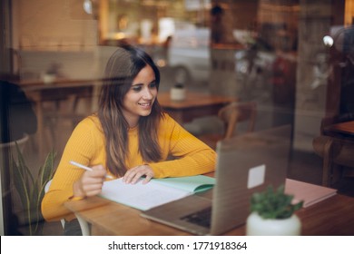 Young Female Student Writing Essay On Modern Topic Using Laptop Computer In Cozy Coffee Shop. Attractive Girl With Netbook Making Notes In Notebook In Public Area