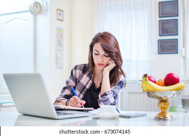 Young Female Student Writing In Workbook And Using Laptop At Home.