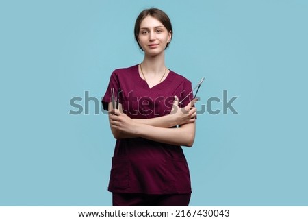 Young Female Surgeon Posing with Steel Surgical Instruments on blue background studio. Woman Doctor Medical Employee wears Medical Uniform Surgical Suit. Concept Training Intern in Practice