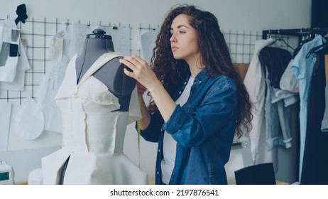 Young female tailor is adjusting clothes on tailoring dummy with sewing pins and measuring with measure-tape. Women's garments, sketches on wall, tailoring items and tools are visible. - Shutterstock ID 2197876541