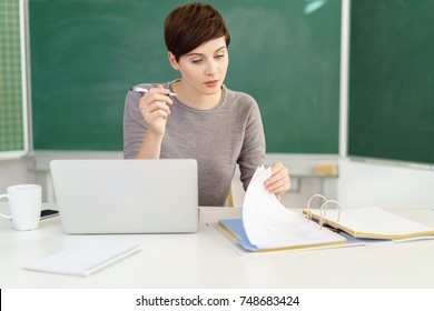 Young female teacher browsing files while sitting at desk with laptop in classroom
