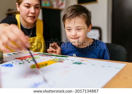 Young female teacher explain, talk at desk with a down syndrome schoolboy. Color painting for disabled kids, child who are down syndrome and student teacher, development of children with spesial needs