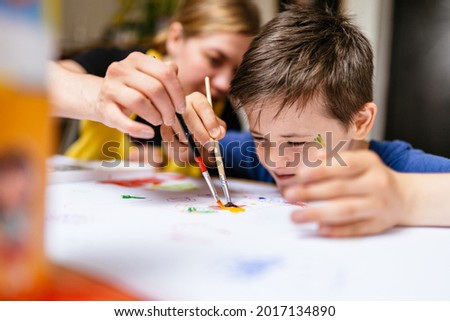 Young female teacher explain, talk at desk with a down syndrome schoolboy. Color painting for disabled kids, child who are down syndrome and student teacher, development of children with spesial needs