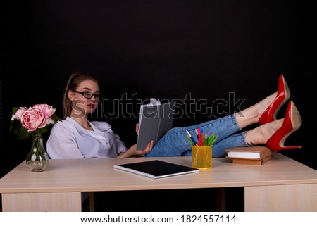 Young female teacher with her feets on the table. Attractive young teacher in red shoes on black background.