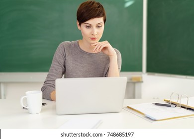 Young female teacher sitting at desk in front of laptop in classroom