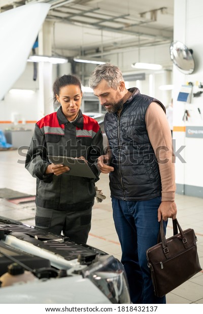 Young female technician of car repair service\
pointing at document while standing by open engine compartment and\
consulting male client