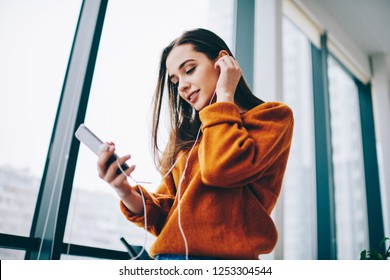 Young female teenager enjoying favourite music playlist listening via application on cellular gadget and electronic earphones, hipster girl spending time for audio book while installing new media app - Shutterstock ID 1253304544