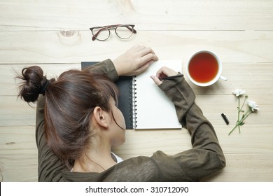 Young Female Teenager Writing Or Drawing On Notebook With Cup Of Tea And White Flowers On Wooden Table. Top View