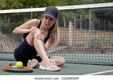 Young Female Tennis Player Sitting On Court Injured Knee While Playing A Match And Showing A Face Of Pain