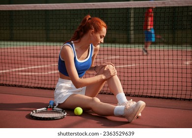 Young female tennis player sitting on court injured knee while playing a match and showing a face of pain - Powered by Shutterstock