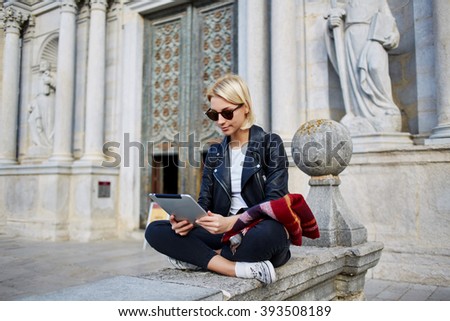 Young female tourist in sunglasses is searching way in navigation on touch pad, while resting after tour in old church. Woman is enjoying good movie on digital tablet during her free time in weekend