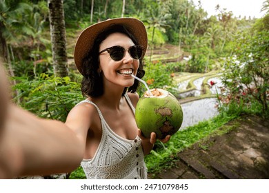 Young female tourist taking selfie with phone while drinking coconut water in Tegalalang rice terrace, Bali. Copy space. Vlogging and vacation concepts. - Powered by Shutterstock