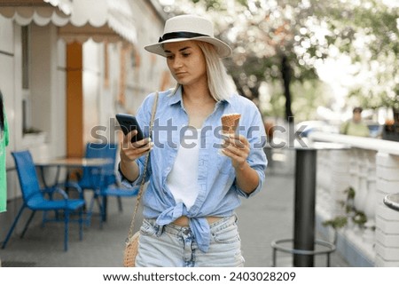 Young female tourist walking through the old town eating ice cream and using a smartphone to search for a map online. Copy space