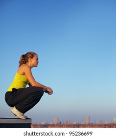 Young Female Traceur Balancing On The Edge Of A High Industrial Urban Building In Readiness For Parkour.