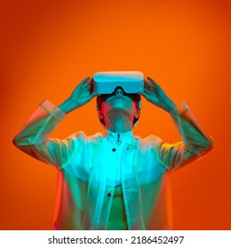 Young Female In Transparent Futuristic Coat Touching VR Headset And Looking Up While Exploring Cyberspace Under Neon Light Against Orange Background.