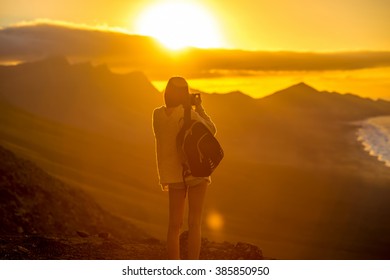 Young female traveler with backpack and photo camera photographing beautiful Cofete coastline with mountains on the sunset on Fuerteventura island in Spain.  - Shutterstock ID 385850950