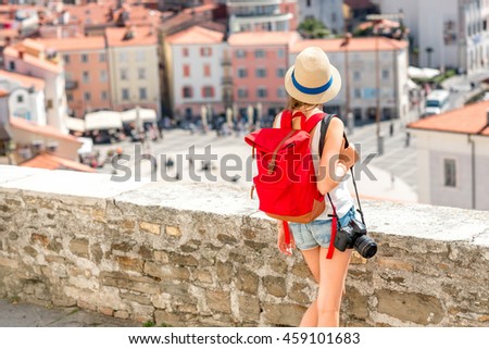 Young female traveler with hat, red backpack and photo camera enjoying the view on Piran old town. Back view with copy space. Traveling in Slovenia