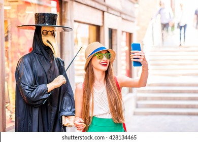 Young Female Traveler Making Selfie Photo With Traditional Venice Doctor Manikin With Mask And Black Costume On The Street In Venice