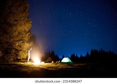 A young female traveler sits by the campfire near her tent, enjoying a moment of relaxation beneath the beautifully starry sky. As the crackling flames provide warmth and comfort - Powered by Shutterstock