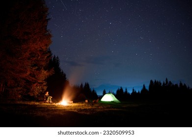 A young female traveler sits by the campfire near her tent, enjoying a moment of relaxation beneath the beautifully starry sky. As the crackling flames provide warmth and comfort - Powered by Shutterstock