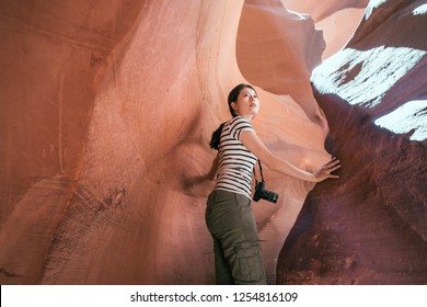 Young Female Traveler Touching The Rock Turning Back Her Head While Hiking In Upper Antelope Canyon. Tourist With Camera Carefully Climbing The Desert Cave On Sunny Day. Asian Girl Love Travel Wild.